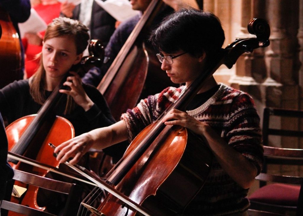 A woman with short dark hair and glasses wearing a jumper, playing the cello.