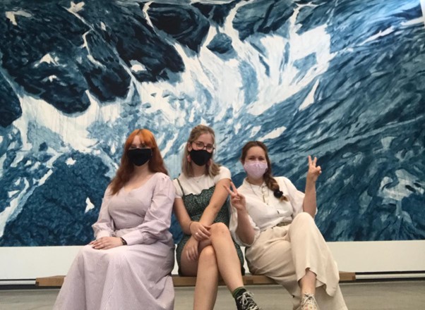 Students sat on a bench wearing face masks with an Exhibition artwork behind them