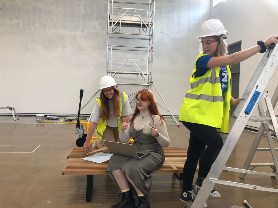 Two women in high vis, one on a stepladder, and a third sat with a laptop