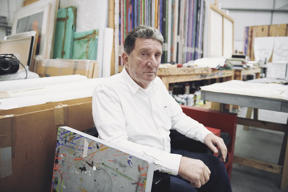 An older man with short grey and white hair looking into the camera as he sits in an art studio, surrounded by supplies and art.