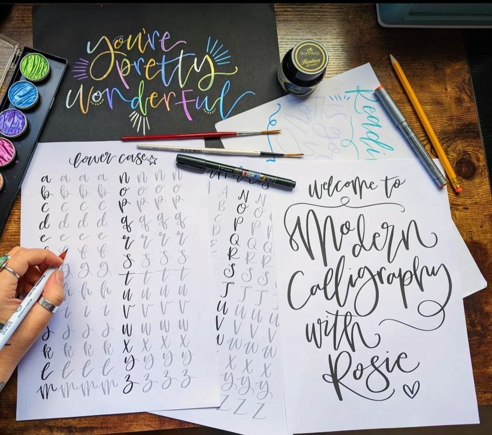 A quote in modern calligraphy and practice sheets laid out on a table with various writing stationery and paints surrounding them.