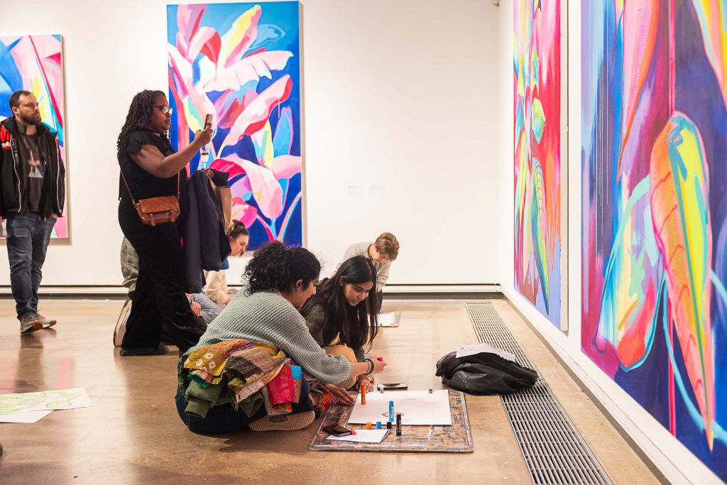 A group of people sat on the floor drawing in front of artwork in a gallery, with people behind standing and taking pictures.