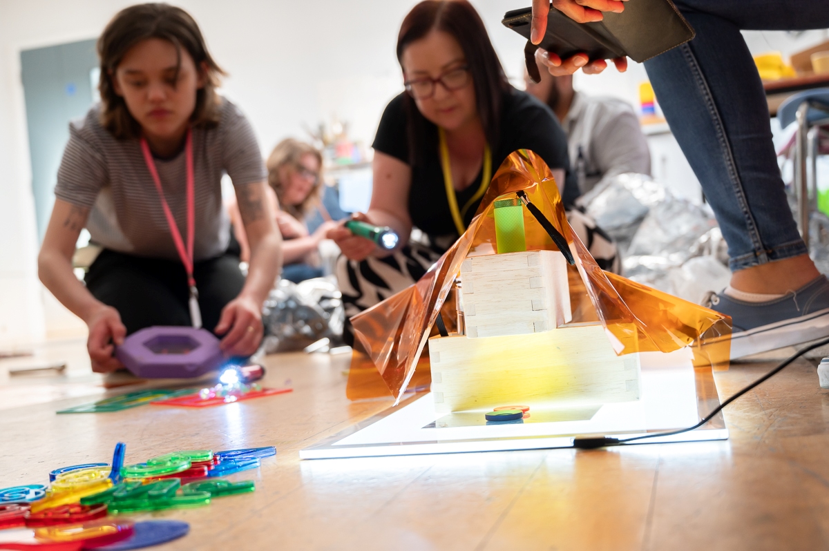 A stack of wooden blocks with yellow cellophane over the top, sitting on a lightbox. To women in the background are shining lights and lifting up shapes.