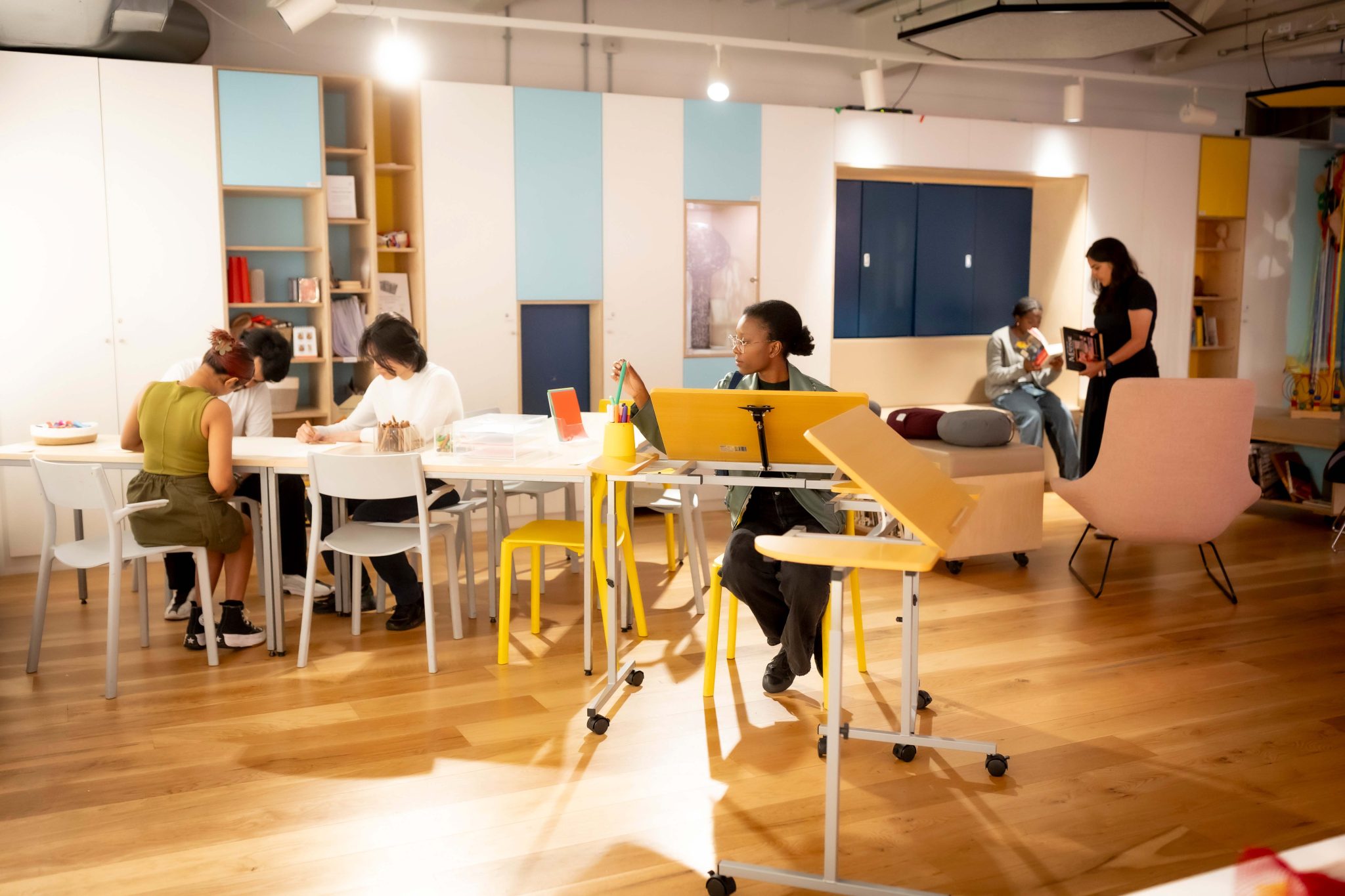 A creative community space in an gallery, with people sat around drawing and creating together.