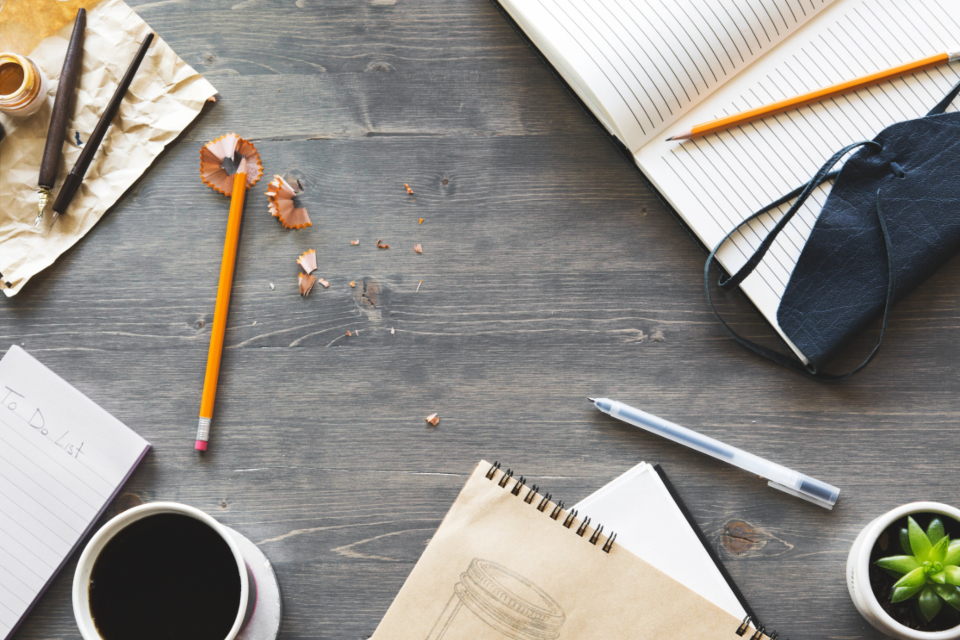 A desk with pens, paper, coffee and notepads scattered around.