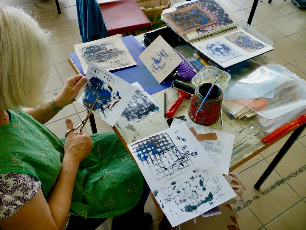 A women with lots of art materials on a table, cutting out paper with ink blotches on it.
