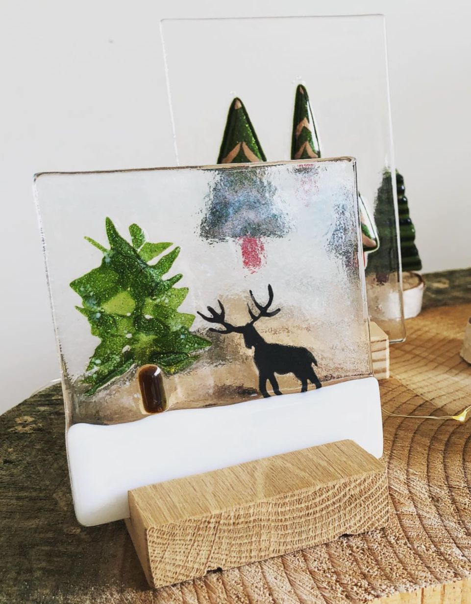 A glass scene of snow, a Christmas tree and a reindeer on a wooden plinth.