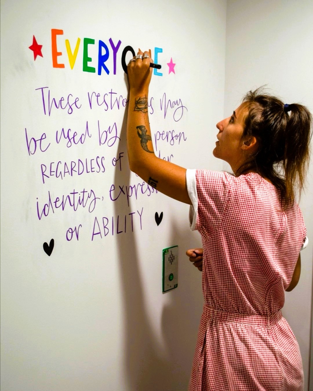 A white women in a red dress drawing colourful words onto a white wall.