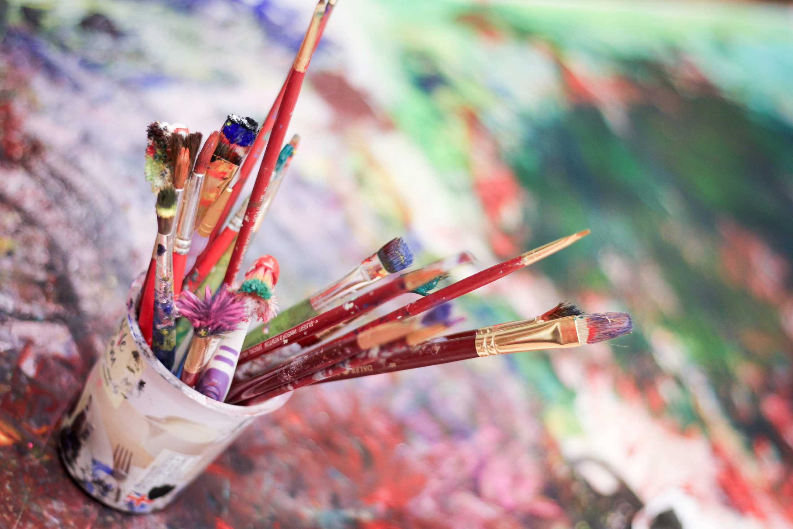 Used, colourful brushes in a pot on a painted, blurry surface.