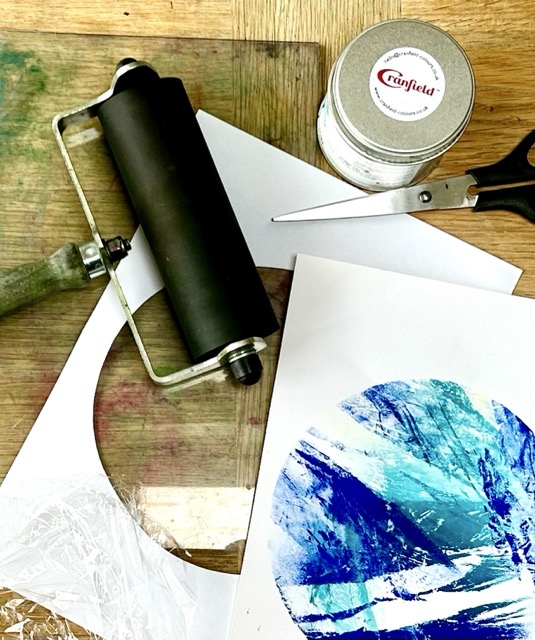 A roller and inks are scattered around paper with coloured prints.