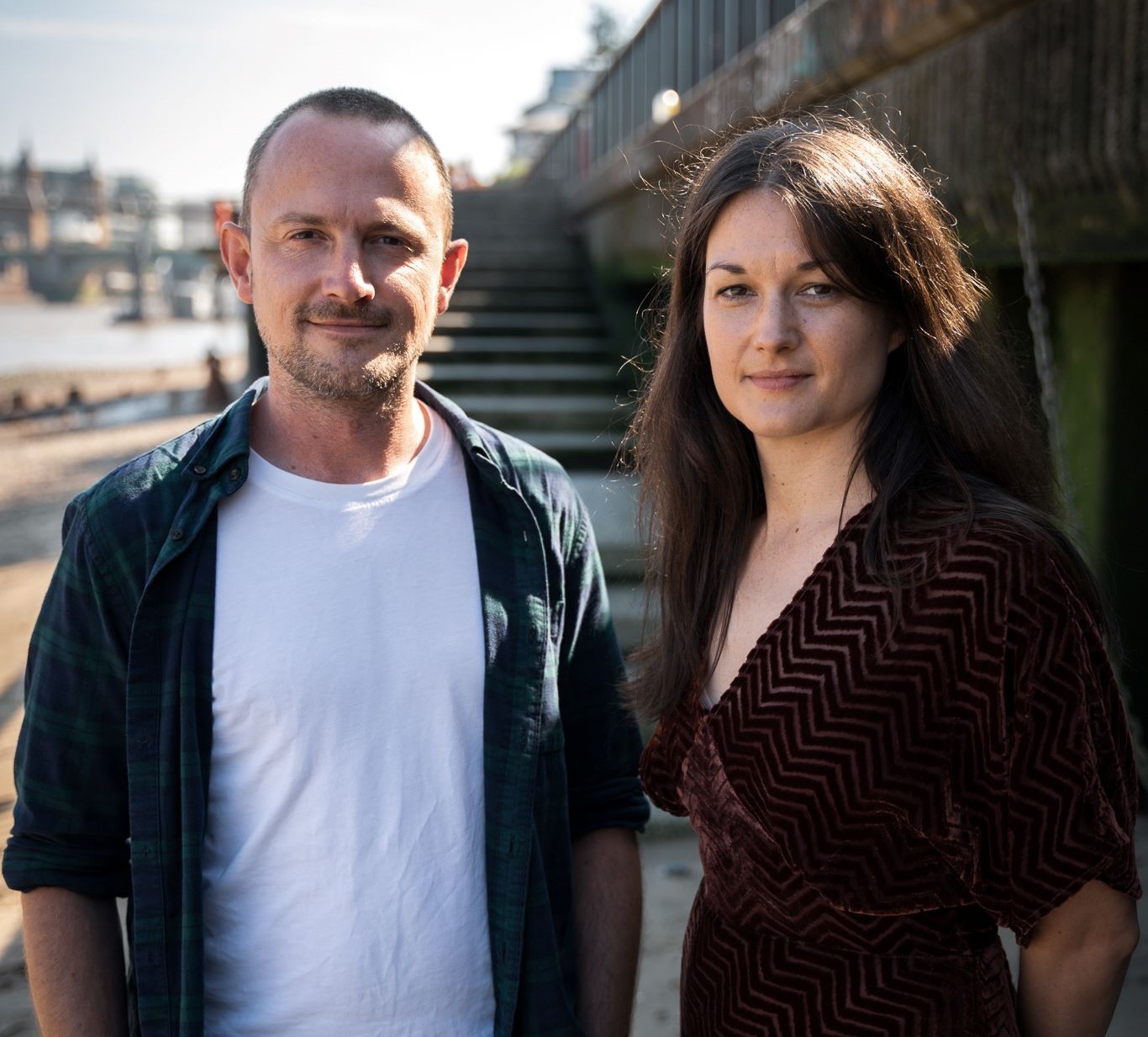 A short haired white man and a long-haired brunette white women stood on a beach together, smiling at the camera.