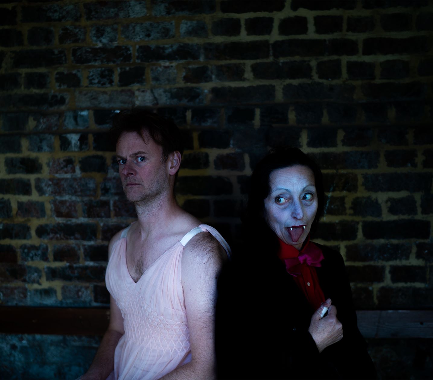 A white man looking worried in a pink dress, next to a white women with vampire teeth and her tongue our looking at him.