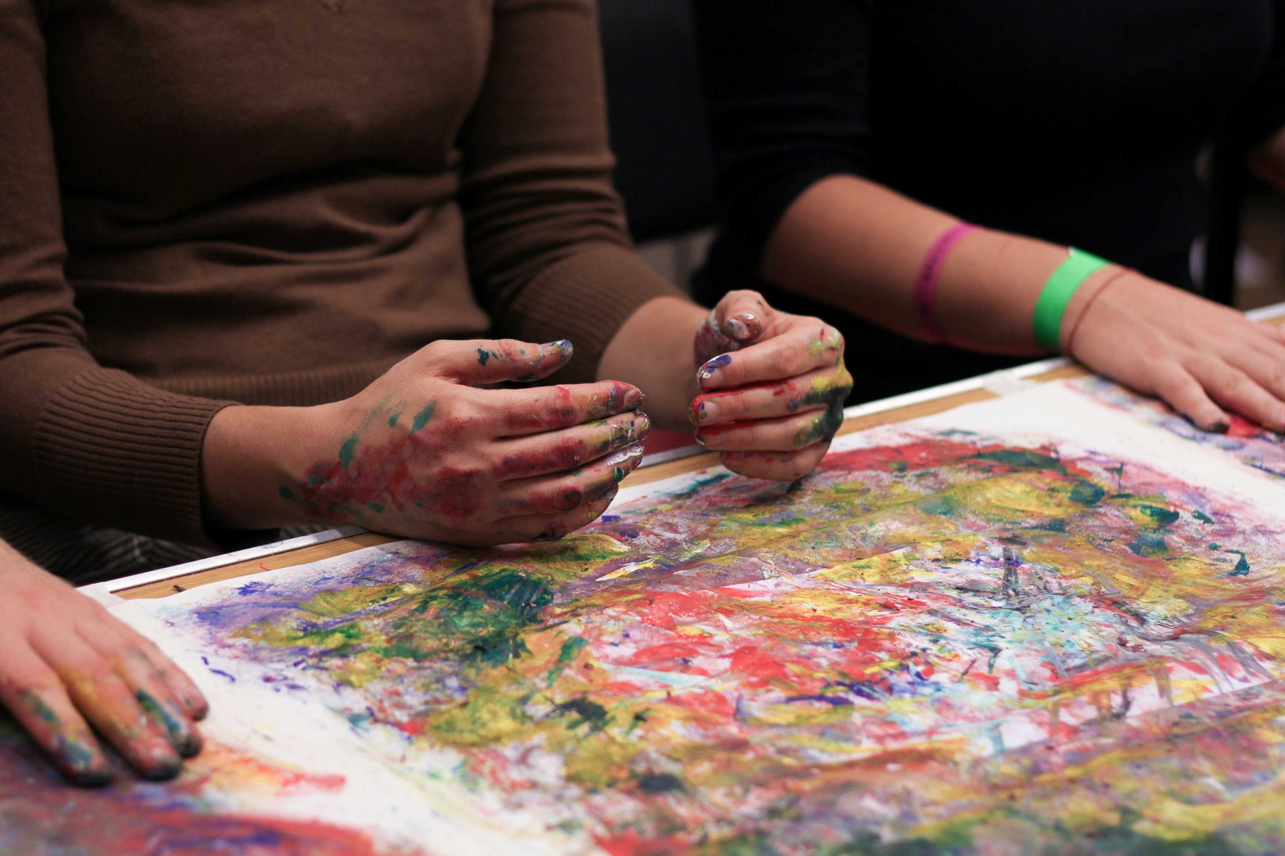 Two hands covered in paint laying in front of a piece of paper covered in many colours of swirled paint.